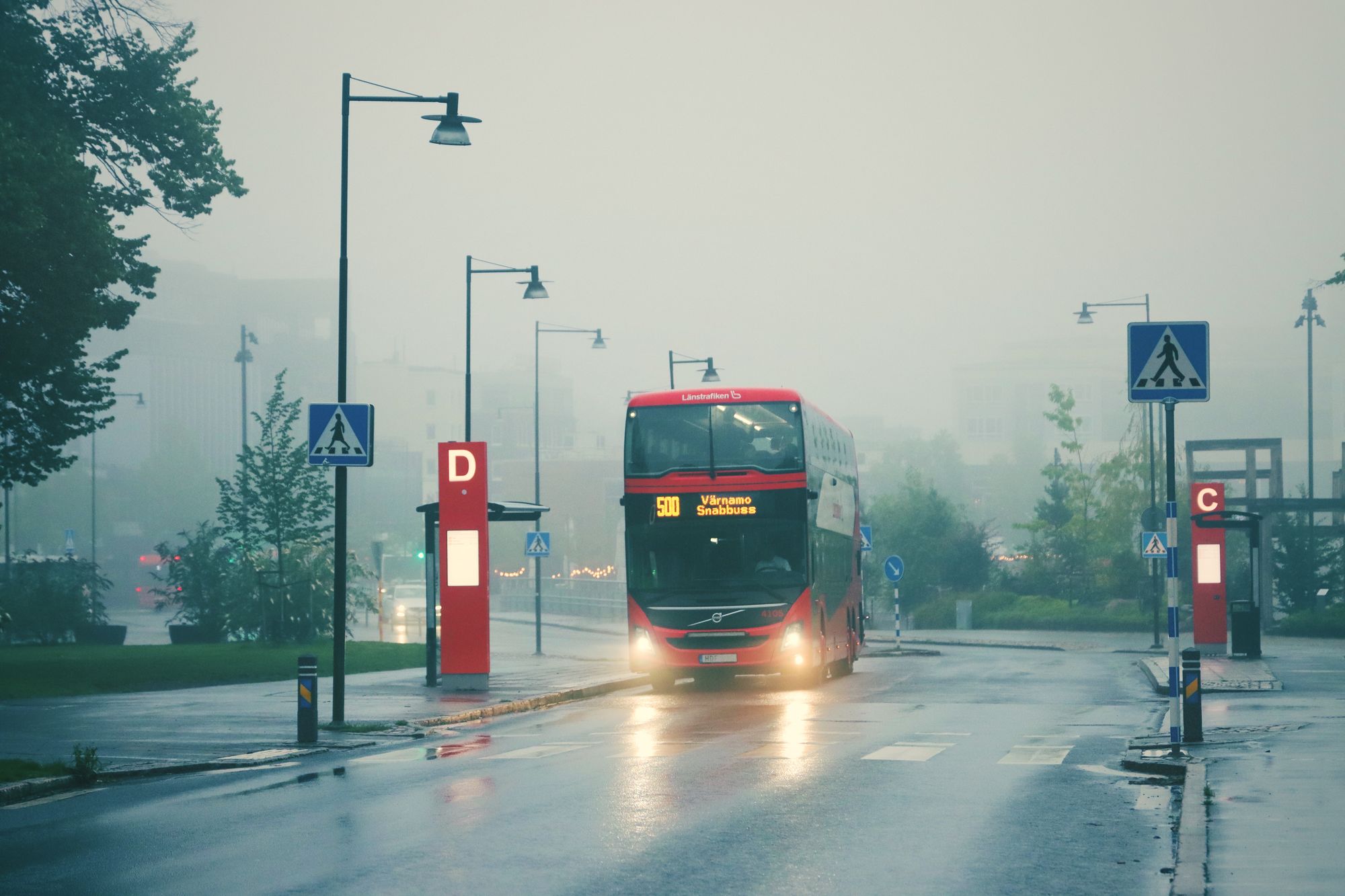 Röd Länstrafiken-buss med tända strålkastare kör in mot en hållplats i dimma